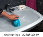 Small photo of White ceramic sink and plunger in female hand clears blockage. Plumbing tool for mechanical cleaning of blockages in sewer pipes and removal of mud from them, preventing the movement of water.