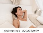 woman wake up in morning in boho style bedroom, sitting on bed, listening to music on earpods, relaxing