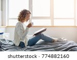 attractive woman sitting on bed in the morning, drinking tea, reading book, curly hair, casual style, blue jeans, white sweater, feeling comfortable at home, having rest, smiling
