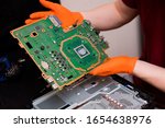 Small photo of SAINT-PETERSBURG, RUSSIA, OCTOBER 27 2019: Man in orange gloves parses the PlayStation 4 Pro console. Close-up, shallow depth of field