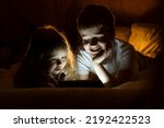 Two children brother and sister having fun, lying comfortably on the bed with great interest watch cartoons on notebook at night at home