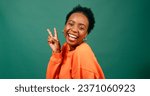 Small photo of Happy young woman holds peace sign to eye, silly pose, green studio portrait