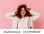 Portrait of charming lady with red lipstick and snow-white smile dressed in bright eco-coat and touching her curly hair on pink background