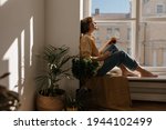 Lifestyle portrait of young brunette sitting on windowsill in light room. Cute short-haired lady wearing orange shirt and blue jeans resting, holding cup of coffee and phone near green