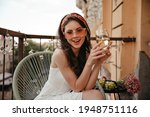 Pretty lady in white dress and sunglasses sits on terrace and holds white wine glass. Young woman with dark hair in red bandage on her head smiles