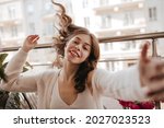 Small photo of Adolescent belarusian female playing with brunette hair softly and touching lens. Enjoying summer time on the daylight veranda in genteel white clothes next to bouquet of flowers