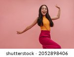Cheerful asian girl with loose dark hair having fun and posing on pink background. High quality photo of young beauty dancing in studio, in summer colorful clothes.
