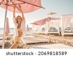 Small photo of Beautiful caucasian lady stretches arms upstairs looking to side on deserted beach. Blonde with bandage on her head in separate swimsuit and shirt on top. Rest and recovery concept.