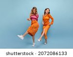 Small photo of Full-length two beautiful cheerful caucasian young girls are dancing in photo studio. Blonde in pink top, skirt. Brown-haired woman in orange dress with bare shoulder in shoes.