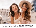 Small photo of Close-up photo of two young fair-skinned girls taking photo against blurred sky background. Fair-haired lady with transparent glasses and hat on her head. Brunette in white top with red necklace.