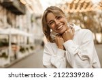 Close up portrait caucasian young happy woman with fresh and clean skin stands outside. Smiling blonde holds collar of white sweatshirt. Lifestyle, female beauty concept
