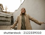 Small photo of Happy young light-skinned woman smiling with teeth outdoors and walking along city street. Blonde in grey sweater and beige cuff. Emotions and states of mind, concept.