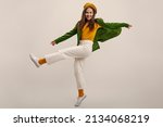 Full length young caucasian girl moves vigorously waving legs on white background in studio. Brunette wears yellow beret, blouse, green jacket and pants. Good mood, fashion trends