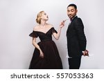 Small photo of Gorgeous young woman in evening dress with flower looking impishly at handsome man in tuxedo with present behind his back on white background. Playful lovely couple, Valentine`s day, surprise
