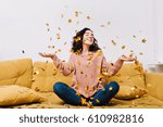 Small photo of Expressing true positive emotions of young joyful woman with cut curly hair having fun in falling tinsels on couch in modern apartment. Home cosines, joy, smiling