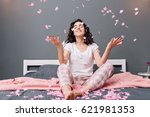 Small photo of Expressing true positive emotions of young joyful woman in pajamas with cut curly hair having fun in falling pink tinsels on bed in modern apartment. Home cosines, smiling with closed eyes