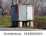 Old dilapidated partially rusted metal shipping container converted into living area with metal doors and two windows on one side put on six concrete pillars foundation and left near paved road