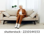 Small photo of a sweet, amiable elderly woman in a brown suit is sitting on a beige wide sofa, smiling pleasantly while in the comfortable environment of her apartment