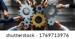 Small photo of Group of business people joining together silver and golden colored gears on table at workplace top view