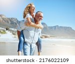 Happy mature couple enjoying vacation by the beach. Active senior husband giving his wife a piggyback ride while enjoying a sunny day outdoors. Energetic man and woman having fun while on holiday