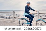 Small photo of Happy, smile and cycling with old woman at beach for fitness, peace and travel with blue sky mockup. Relax, workout and retirement with senior lady riding on bike for nature, health and wellness