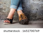 The elegant foot of young teenage girl wearing peep-toe sandals in one foot and hiking boots in another posing against a brick wall wearing blue denim jeans.