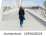 Woman wearing jeans and a warm coat with a large bag over her shoulder walking away across a pedestrian bridge with white receding rails in a town environment
