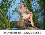 Small photo of Attractive blond woman wearing a gold gogo dress sitting on a tree trunk at the edge of a lake looking aside