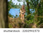 Small photo of Attractive blond woman wearing a gold gogo dress sitting on a tree trunk at the edge of a lake