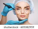 Pretty girl with dark eyebrows wearing blue medical hat at studio background, doctor's hands wearing blue gloves drawing perforation lines on face, plastic surgery concept.