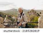 Small photo of Elderly woman smiling at the camera while standing on a hilltop with binoculars. Happy senior woman enjoying a leisurely hike outdoors. Mature woman enjoying recreational activities after retirement.