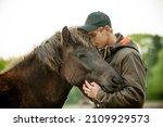Young blonde caucasian man with cap petting a domesticated dark brown Icelandic mare. Human and animal equine friendship. 
