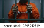 Small photo of Male criminal in orange uniform and handcuffs holds metal bars, stands in prison cell, looks at camera. Prisoner serves imprisonment term in jail. Detention center or correctional facility. Portrait.
