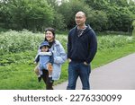Small photo of A multicultural family walking on a footpath in Figgate Park in Edinburgh in spring. The mother is carrying her infant son with a baby carrier. The baby wears a misplaced hat and is uncomfortable.