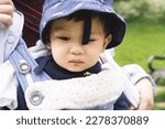 Small photo of Close-up photo of a male infant carried by his unrecognizable parent on a baby carrier wearing a blue misplaced hat. He is sad and looking down, feeling unwell.