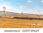 Old Algerian train rolling through plains of golden yellow wheat with a quarry and a pillar of electricity cables, dry tree and blue sky. View from the east-west freeway near the town of Tlemcen. 
