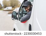 Package delivery and shipping concept. Positive smiling African-American ethnic man in his 30s working as delivery man, looking outside of driver's window at camera. High quality photo