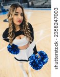Small photo of Cheerleader woman with blue pom pons standing, looking into camera, on a basketball court, high angle view. High quality photo