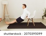 Small photo of Side view of a determined middle-aged man exercising tricep dips or triceps extension with a chair as workout routine for the arms at home during pandemic