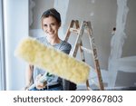 Small photo of Playful young woman doing home improvements laughing as she brandishes a paint roller at the camera