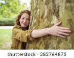 Nature-Lover Blond Teen Girl Hugging Huge Tree Trunk at the Park While Smiling at the Camera.