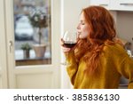Young redhead woman standing in her apartment sipping a glass of red wine with her eyes closed in pleasure, side view