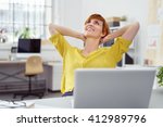 Single daydreaming woman in yellow blouse holding hands behind neck in front of laptop in small office with bright windows