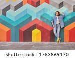 Relaxed young female in denim outfit smiling and looking away while leaning on multicolored graffiti wall with geometric ornament on city street