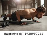 Muscular man doing push up exercise on mat near dumbbells during intense fitness workout in grungy gym