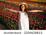 Delighted young female tourist in white dress straw hat and sunglasses, smiling and looking at camera while standing in tulip field during trip in Netherlands