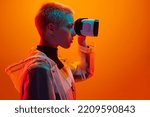 Small photo of Side view of emotionless female experiencing metaverse while using virtual reality helmet on orange background in studio with neon illumination