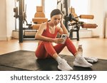 Fit female athlete in sportswear sitting on mat and using fitness app between workout sets in gym