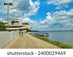 Small photo of Madison, Wisconsin, USA - Aug. 1 2023: Landscape view of bikers on a trail in front of Monona Terrace facing Lake Monona beneath a partly cloudy blue Summer sky in Madison, WI.