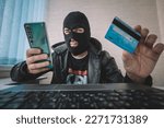 Small photo of Male hacker in a robber mask uses phone, credit card and laptop in some fraudulent scheme. Cyber thief stole the personal data and credit card information. Hacker uses malware to steal user's money.